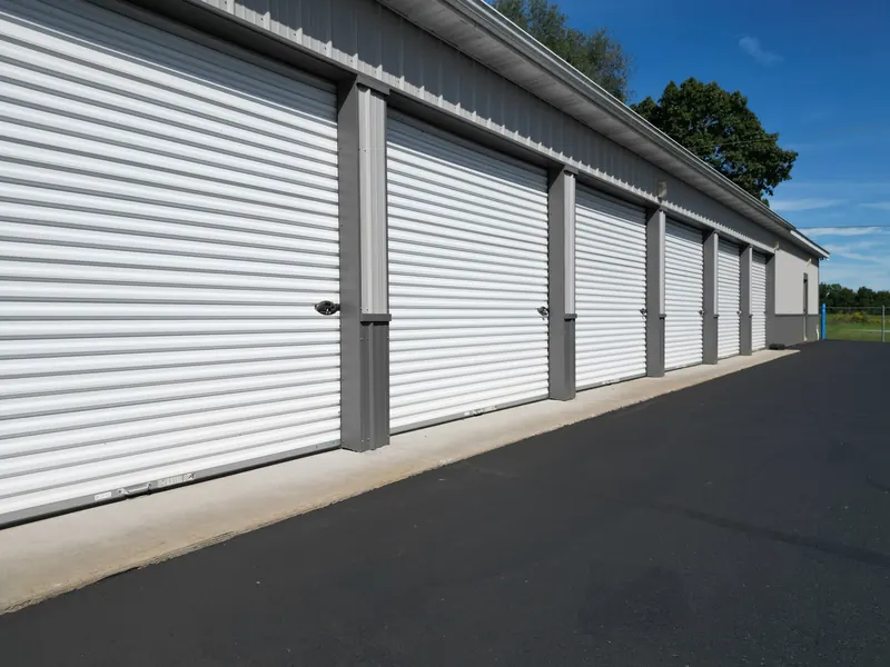 A long row of drive-up storage units with white roll-up doors beside a paved aisle