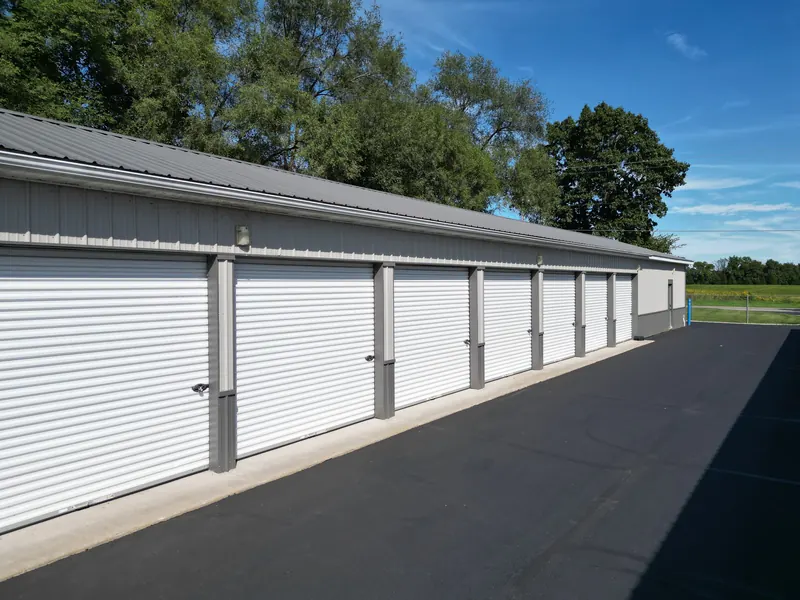 The side of a storage building with a row of roll-up doors and mature trees behind it